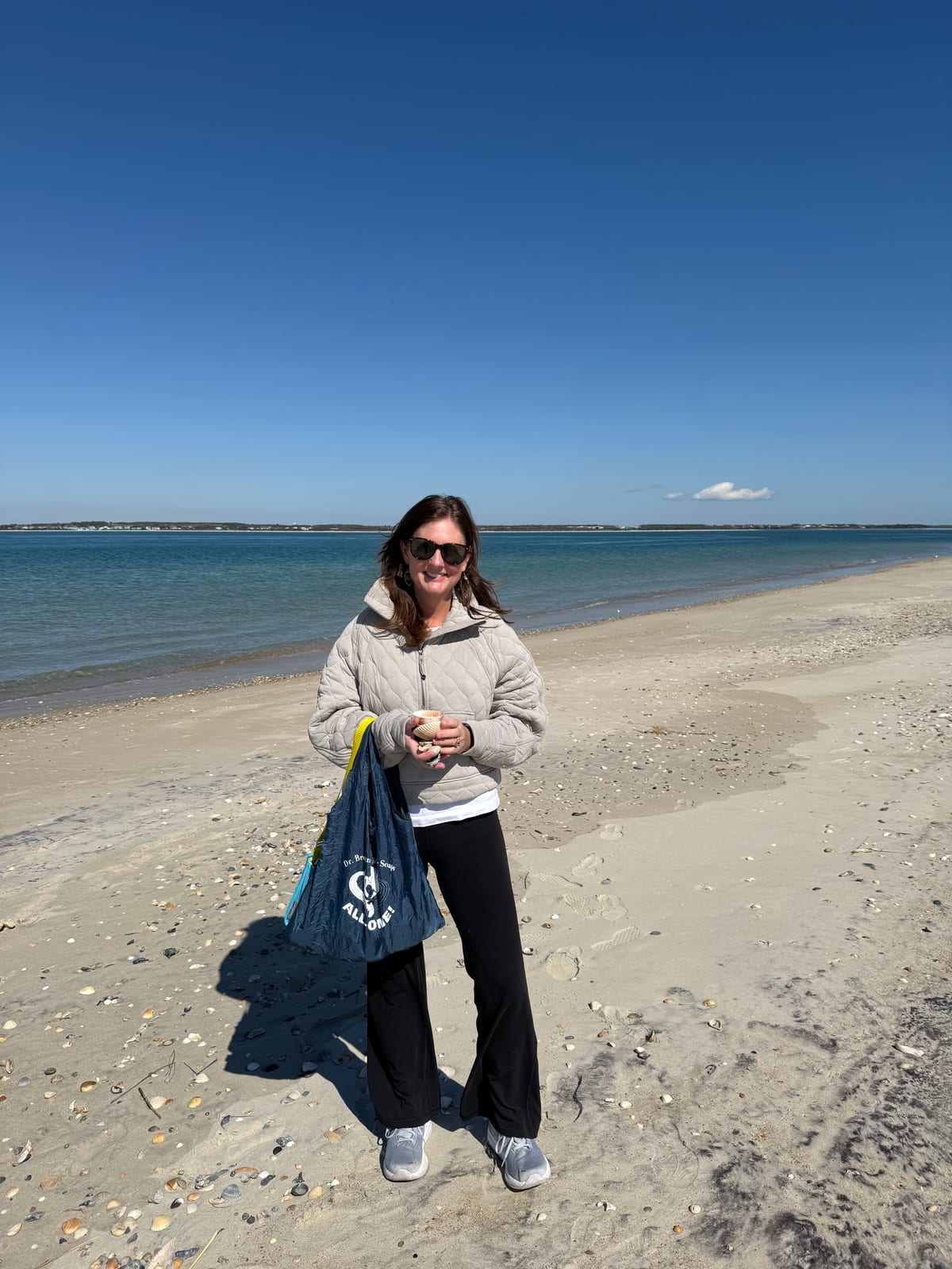 Ali Turner smiling on a Carolina beach, holding a shell, with her shell-seeking bag over her arm