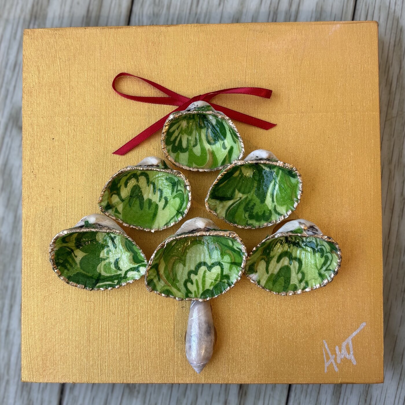 A gold canvas panel with green patterned clam shells arranged into a Christmas tree, topped with a red bow.