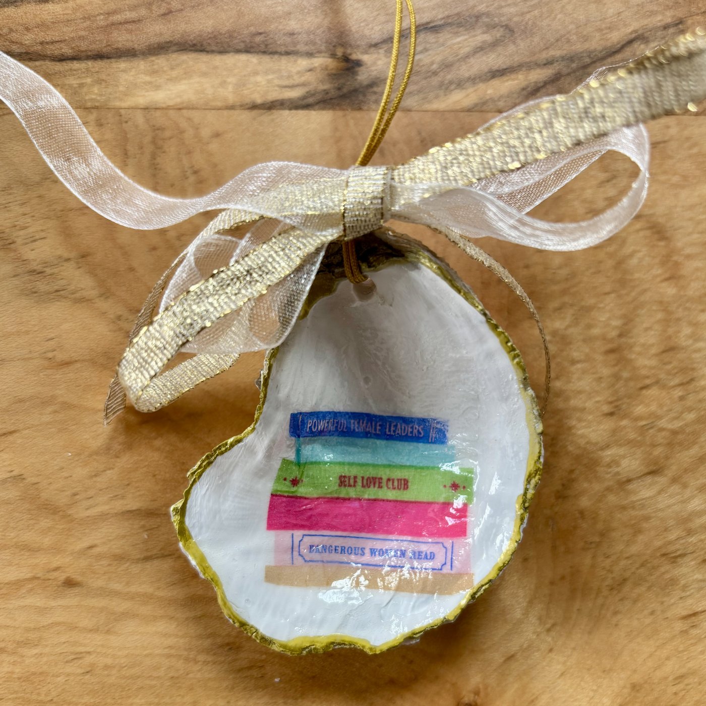 An oyster shell ornament with a gold ribbon bow that illustrates a stack of books, perfect for your book club ladies.