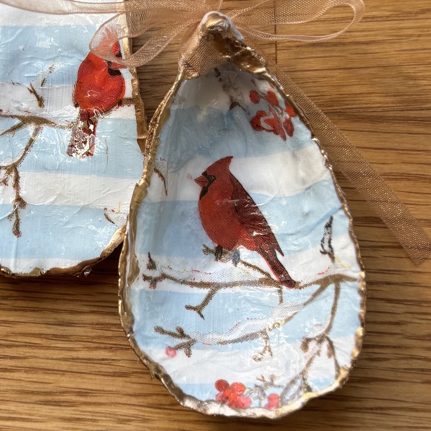 A gold-rimmed oyster shell ornament with a red cardinal on a snowy branch with red berries, over pale blue and white stripes, hung on a peach organza ribbon. Comes in a gift bag.
