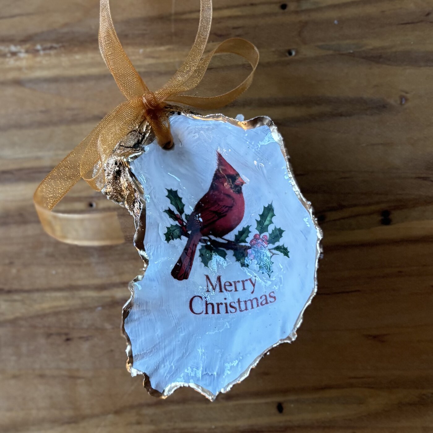 A gold-rimmed oyster shell ornament with a red cardinal on a holly branch and "Merry Christmas" in red, hung on a gold organza ribbon. Comes in a gift bag.