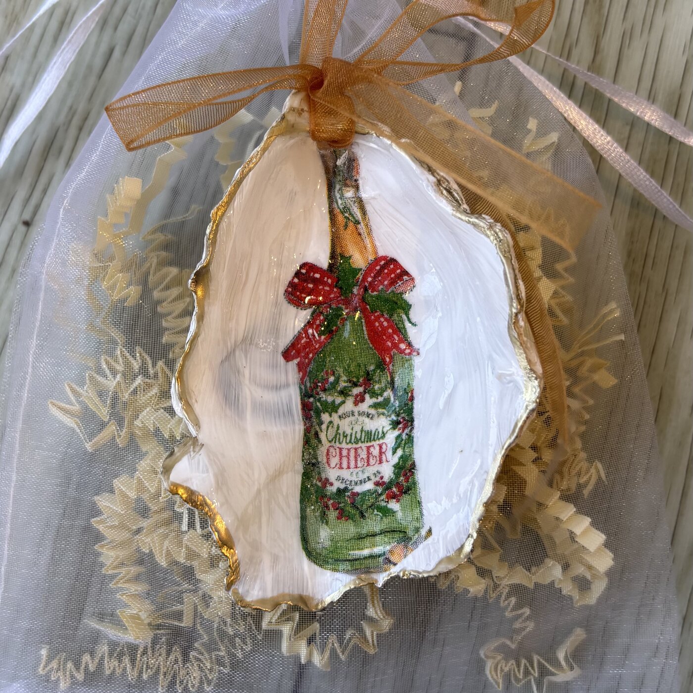 A gold-rimmed oyster shell ornament with a green "Christmas Cheer" bottle tied in a red bow. Comes in a gift bag.