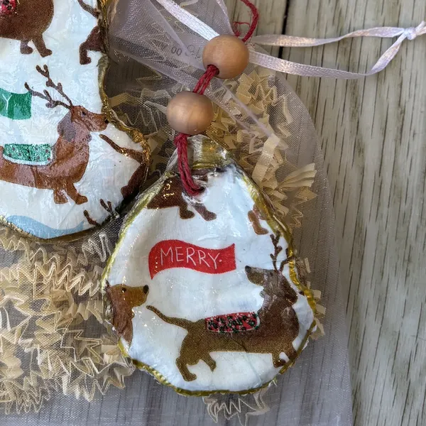 An oyster shell ornament with reindeer-antlered dachshunds and a "Merry" or "Joy" banner. Each one is cut a little differently. Comes in a gift bag.