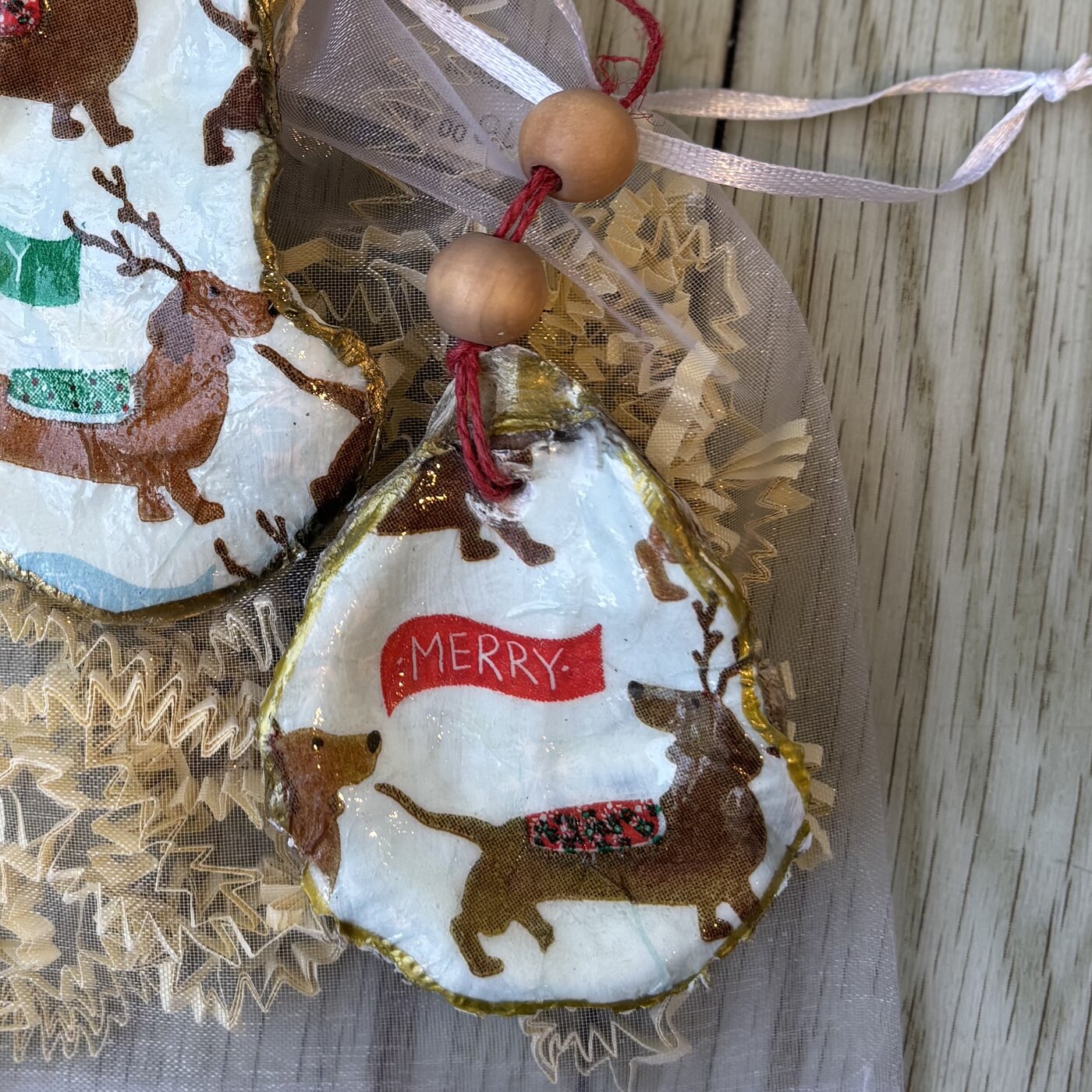 An oyster shell ornament with reindeer-antlered dachshunds and a "Merry" or "Joy" banner. Each one is cut a little differently. Comes in a gift bag.