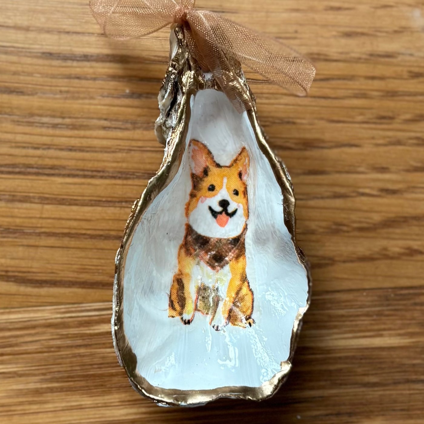 A gold-rimmed oyster shell ornament with a smiling corgi in a plaid bandana, hung on a peach organza ribbon. Comes in a gift bag.