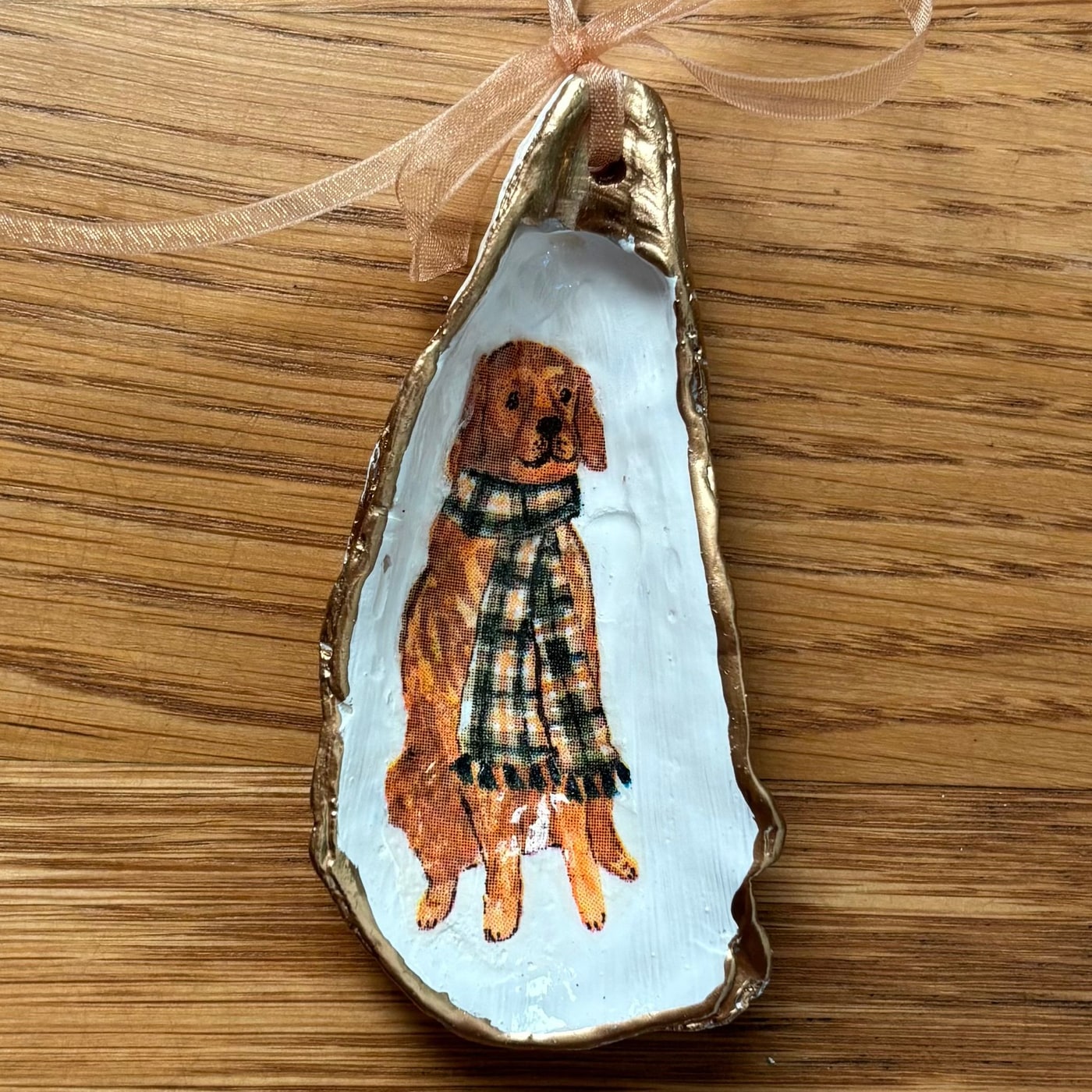 A gold-rimmed oyster shell ornament with a golden retriever in a plaid scarf, hung on a peach organza ribbon. Comes in a gift bag.