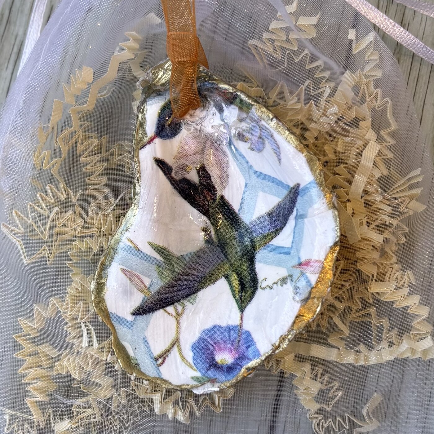 A gold-rimmed oyster shell ornament with a hummingbird and morning glory on a blue trellis. Comes in a gift bag.