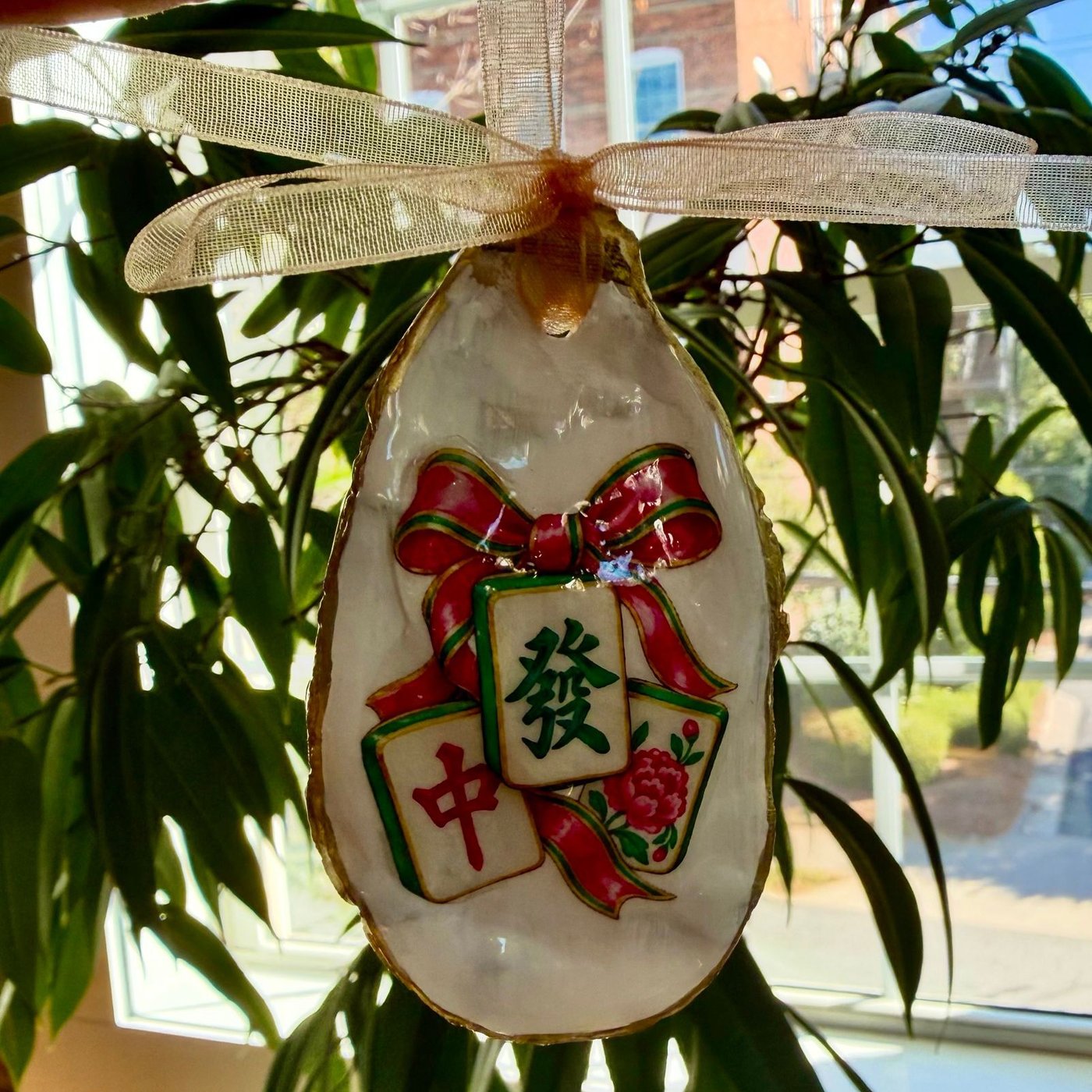 A gold-rimmed oyster shell ornament with three mahjong tiles tied up in a red Christmas bow, hung on a gold organza ribbon. Comes in a gift bag.