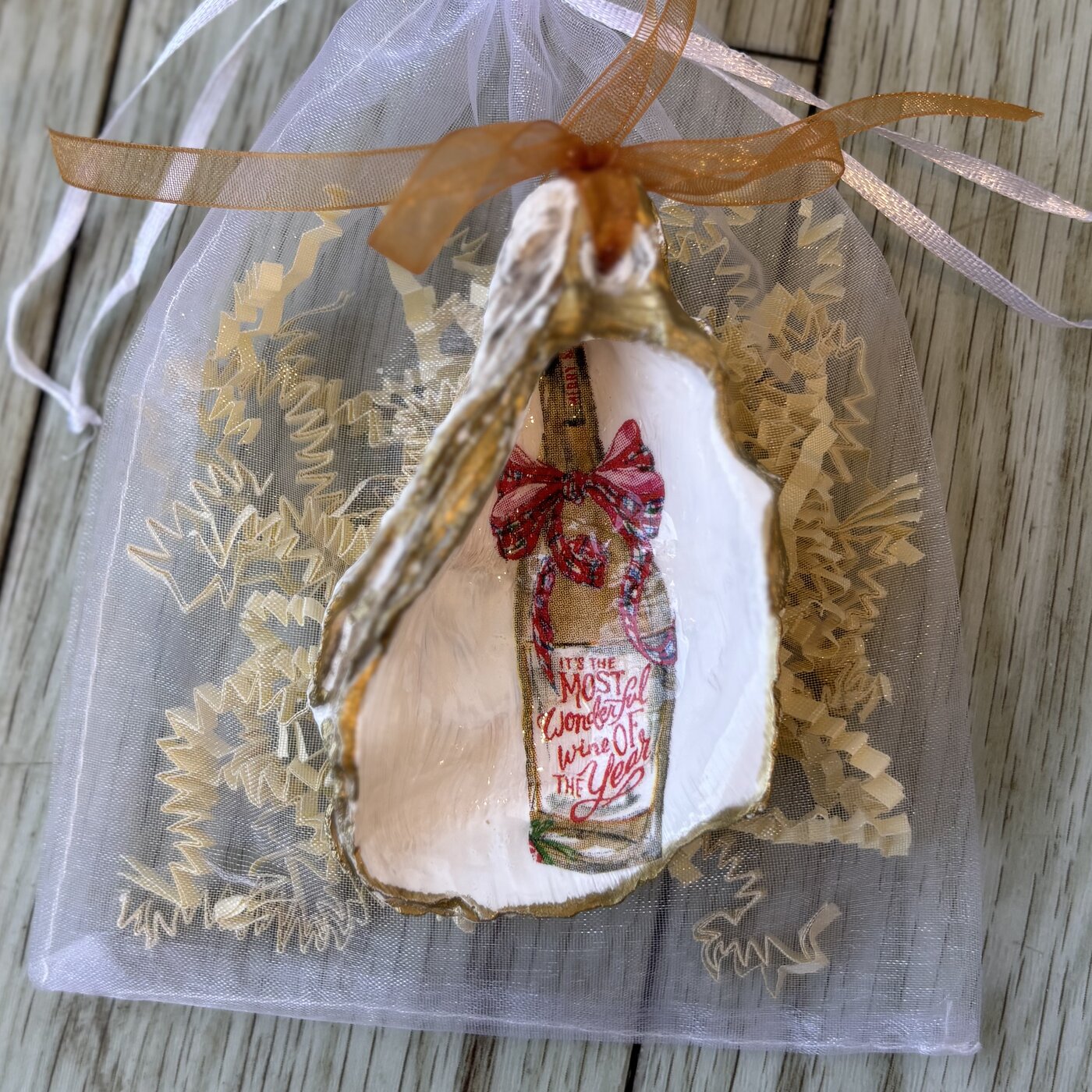 A gold-rimmed oyster shell ornament with a plaid-bowed "It's the Most Wonderful Wine of the Year" bottle. Comes in a gift bag.