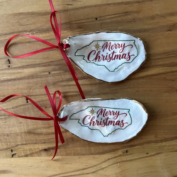 A gold-rimmed oyster shell ornament with the North Carolina state outline, a gold star and "Merry Christmas" in red script, hung on a red ribbon. Comes in a gift bag.