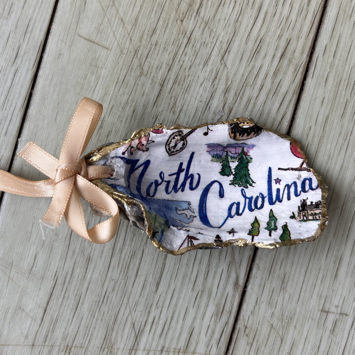 An oyster shell ornament illustrated with the North Carolina state outline and iconic scenery.