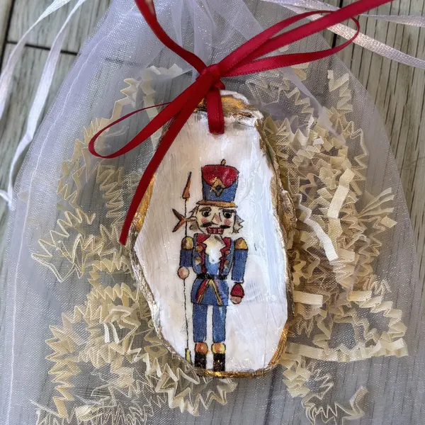A gold-rimmed oyster shell ornament with a classic red and blue nutcracker soldier. Comes in a gift bag.