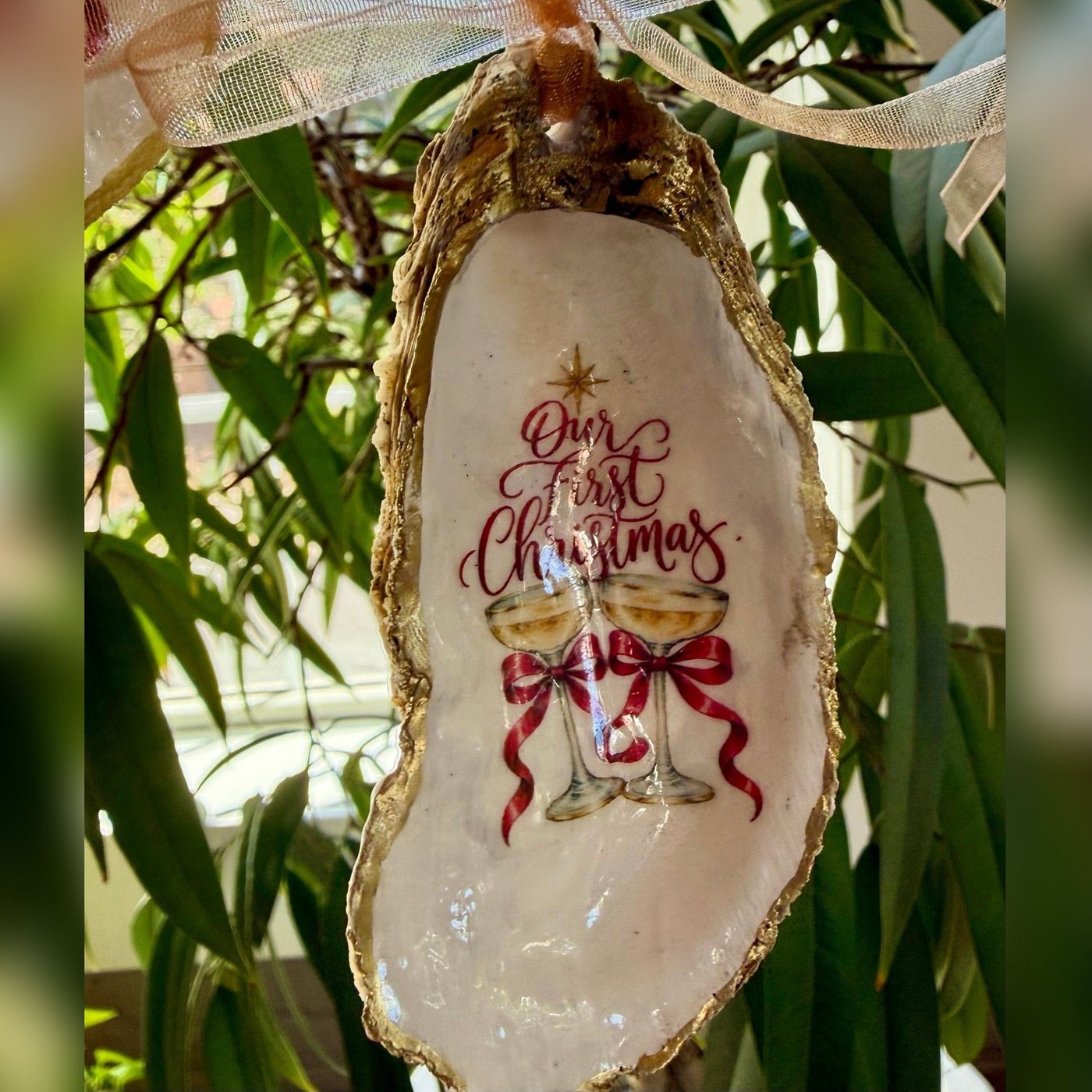 A gold-rimmed oyster shell ornament reading "Our First Christmas" over two champagne coupes tied with red bows, hung on a gold organza ribbon. Comes in a gift bag.