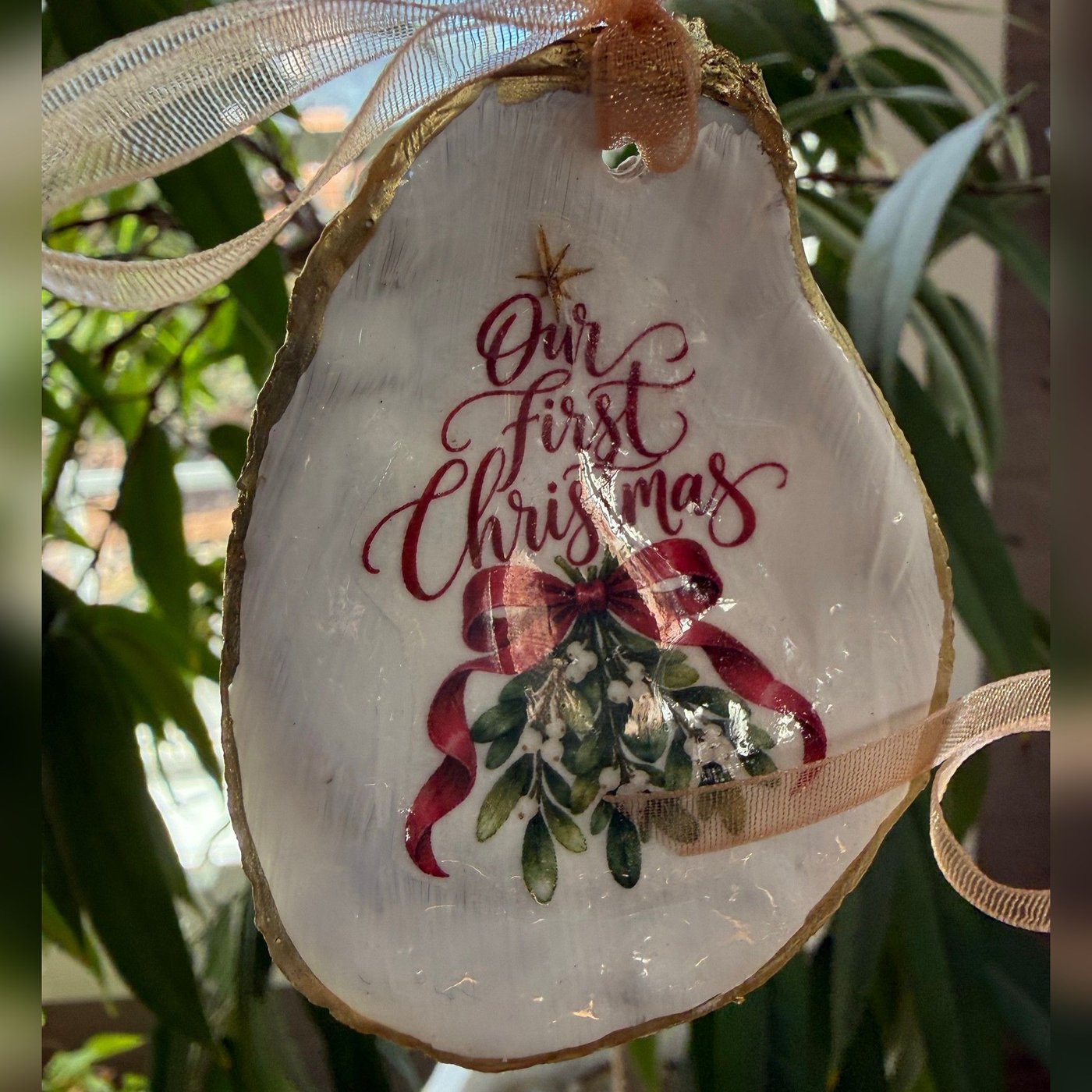 A gold-rimmed oyster shell ornament reading "Our First Christmas" over a sprig of mistletoe tied with a red bow, hung on a gold organza ribbon. Comes in a gift bag.