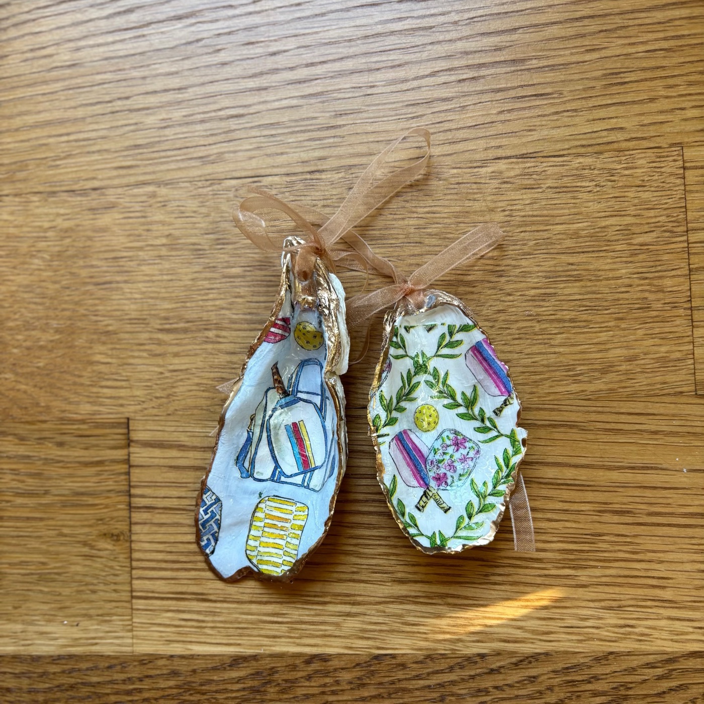 A gold-rimmed oyster shell ornament patterned with pickleball paddles and yellow pickleballs, hung on a peach organza ribbon. Blue has blue lattice and blue-and-yellow striped paddles; Pink has pink-striped and pink floral paddles with green leafy vines. Comes in a gift bag.