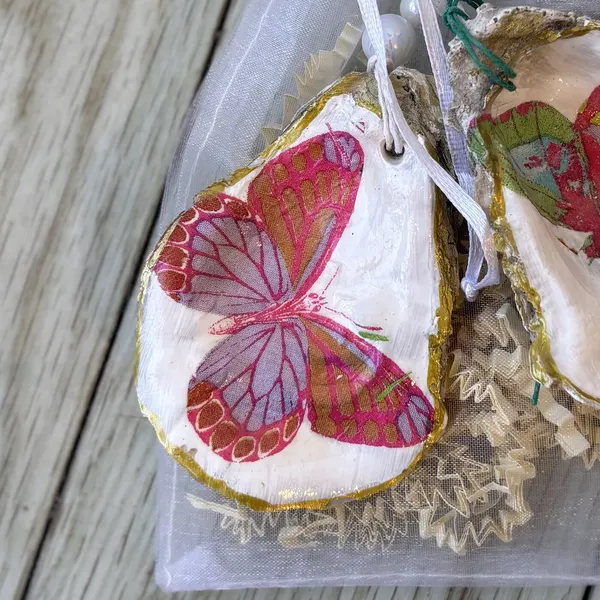 A gold-rimmed oyster shell ornament with a pink and red butterfly. Comes in a gift bag.