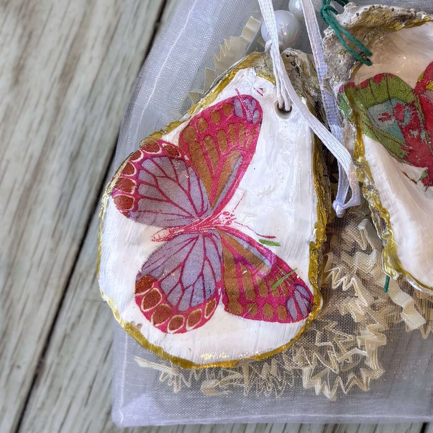 A gold-rimmed oyster shell ornament with a pink and red butterfly. Comes in a gift bag.