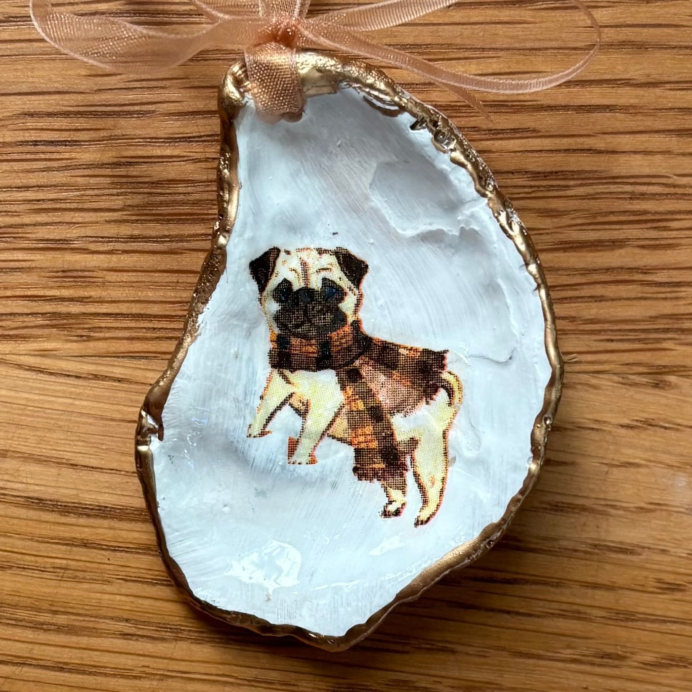A gold-rimmed oyster shell ornament with a pug in a brown plaid scarf, hung on a peach organza ribbon. Comes in a gift bag.