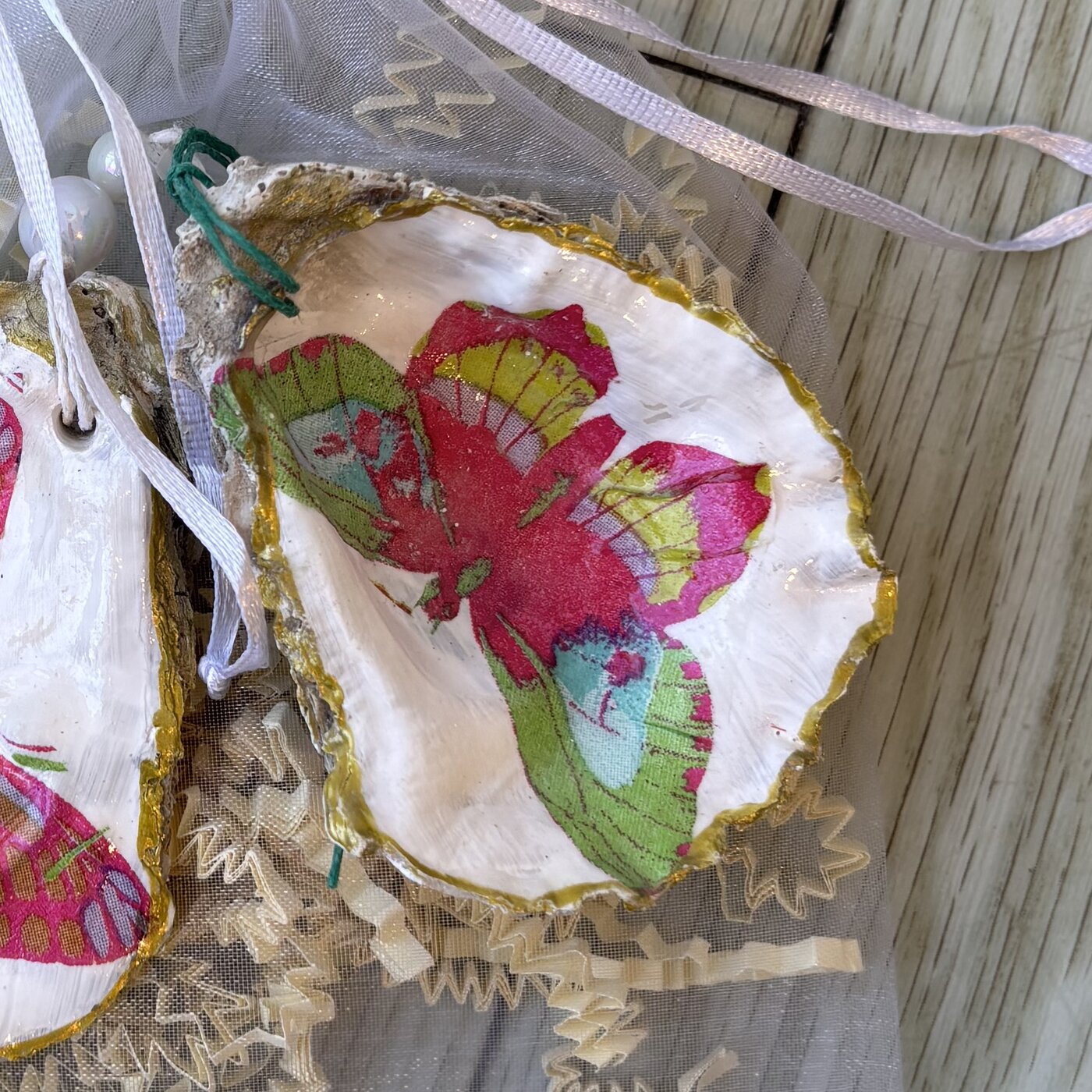 A gold-rimmed oyster shell ornament with a bright pink, green and blue butterfly. Comes in a gift bag.
