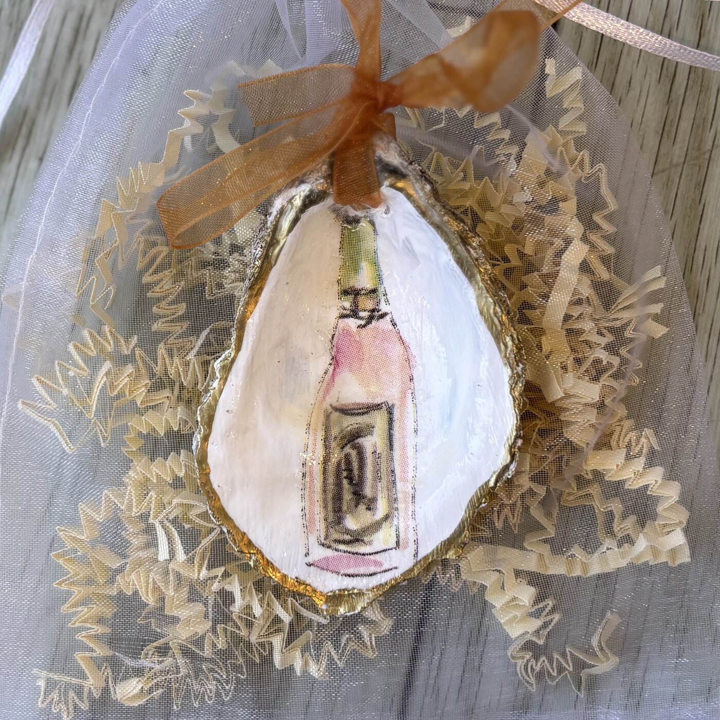 A gold-rimmed oyster shell ornament with a pink rosé bottle design. Comes in a gift bag.