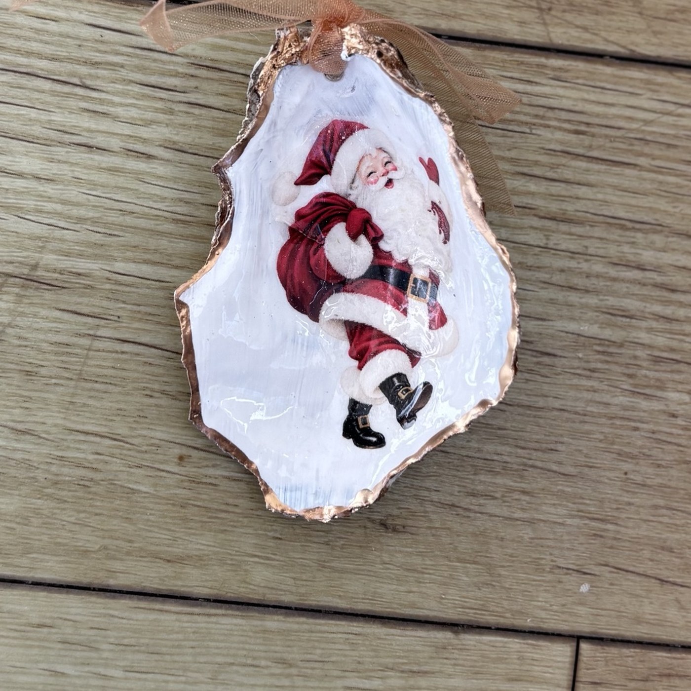 A copper-rimmed oyster shell ornament with a jolly vintage Santa kicking up his heels and carrying a red toy sack, hung on a copper organza ribbon. Comes in a gift bag.