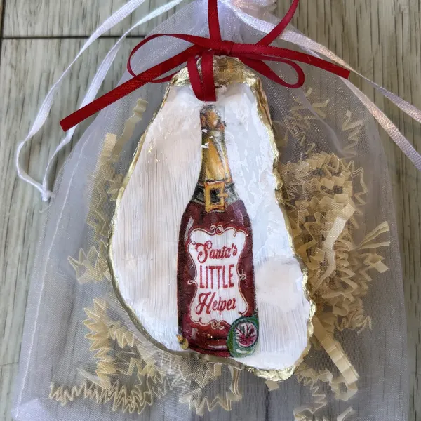 A gold-rimmed oyster shell ornament with a "Santa's Little Helper" bottle illustration. Comes in a gift bag.