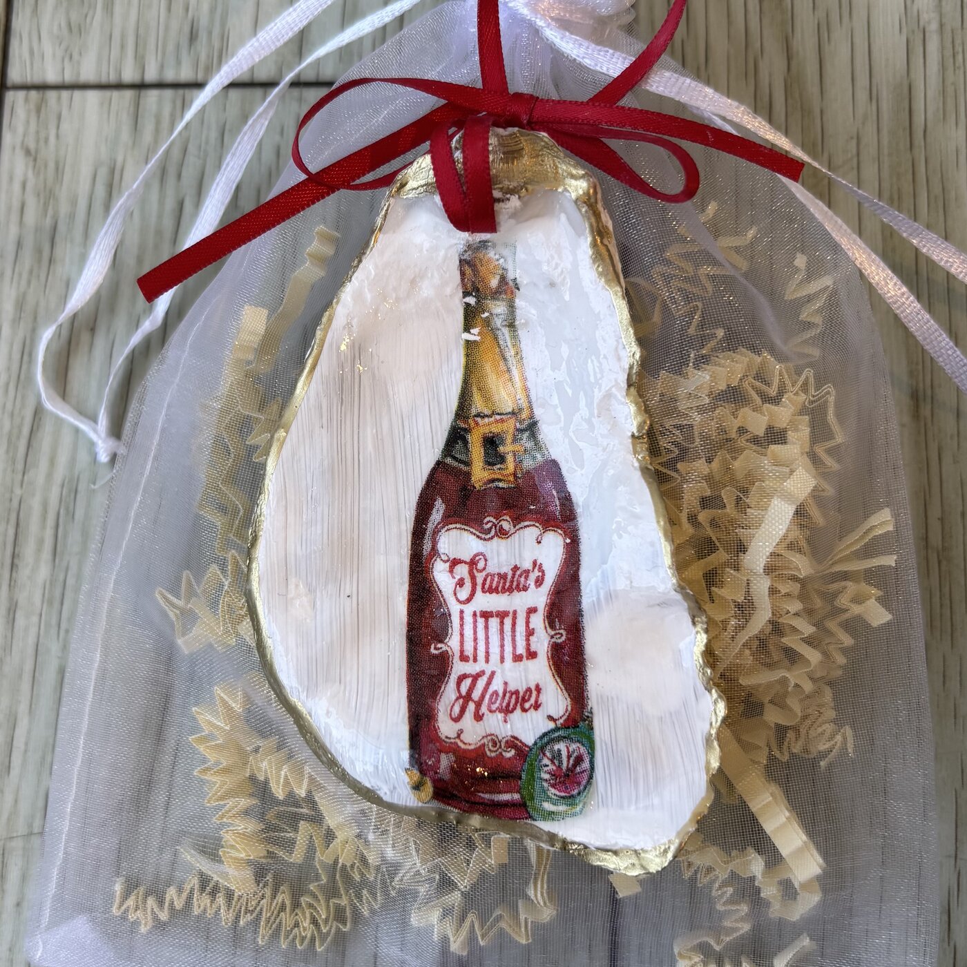 A gold-rimmed oyster shell ornament with a "Santa's Little Helper" bottle illustration. Comes in a gift bag.
