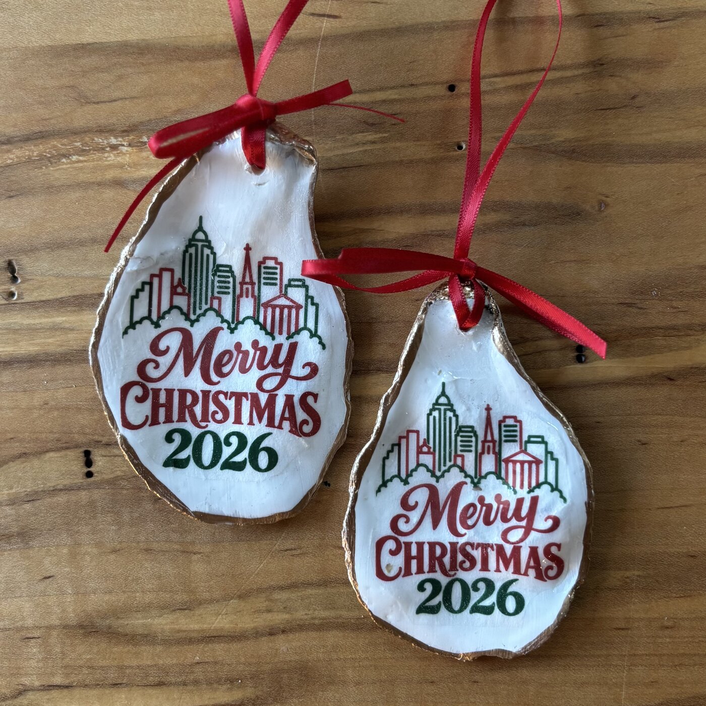 A gold-rimmed oyster shell ornament with the Winston-Salem, North Carolina skyline in green over "Merry Christmas 2026" in red and green, hung on a red ribbon. Comes in a gift bag.
