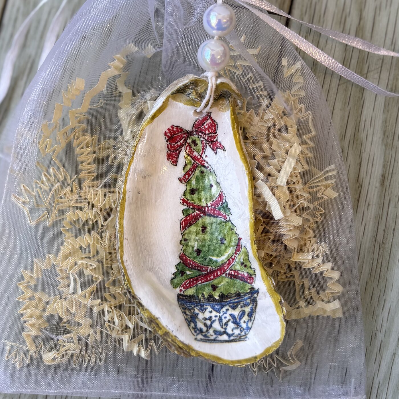 An oyster shell ornament with a ribbon-wrapped topiary tree in a blue and white pot, hung with pearl beads. Comes in a gift bag.
