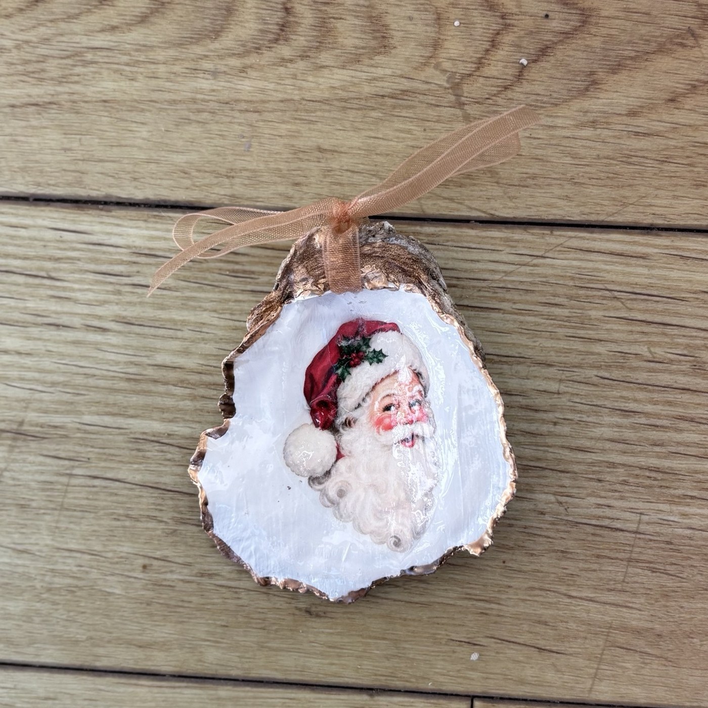 A copper-rimmed oyster shell ornament with a vintage Santa face in a red hat trimmed with holly, hung on a copper organza ribbon. Comes in a gift bag.