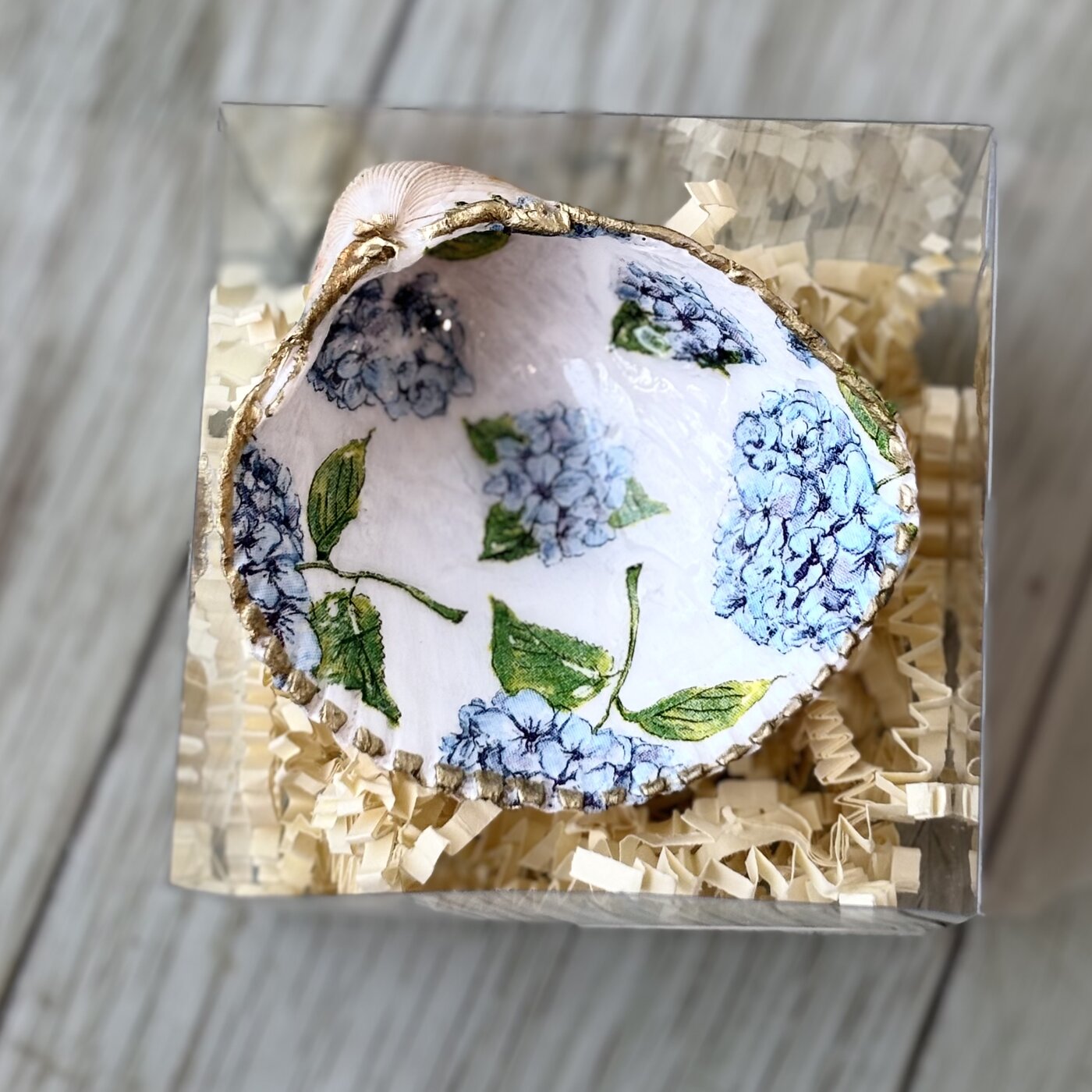 A gold-rimmed shell ring dish scattered with blue hydrangea blooms. Comes in a gift box.