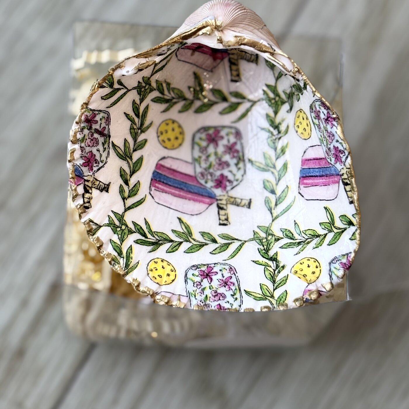 A gold-rimmed shell ring dish patterned with pickleball paddles and balls inside a green laurel wreath. Comes in a gift box.