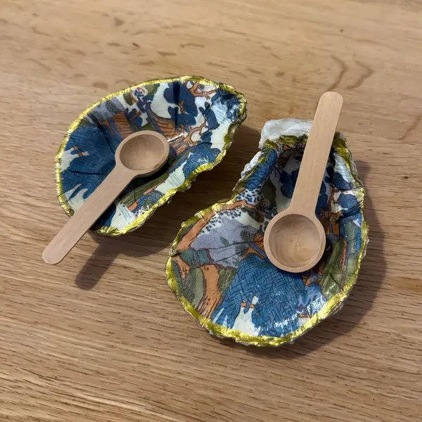 A pair of gold-rimmed oyster shell cellars with a blue and orange chinoiserie pagoda scene, with two gold or wooden spoons. Comes in a gift box.