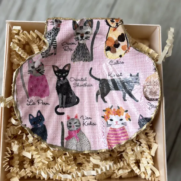 A gold-rimmed shell dish covered in illustrated cats on pink. Comes in a gift box.