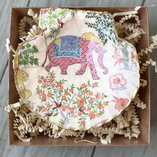 A gold-rimmed shell dish with a pink chinoiserie elephant and a flowering garden. Comes in a gift box.