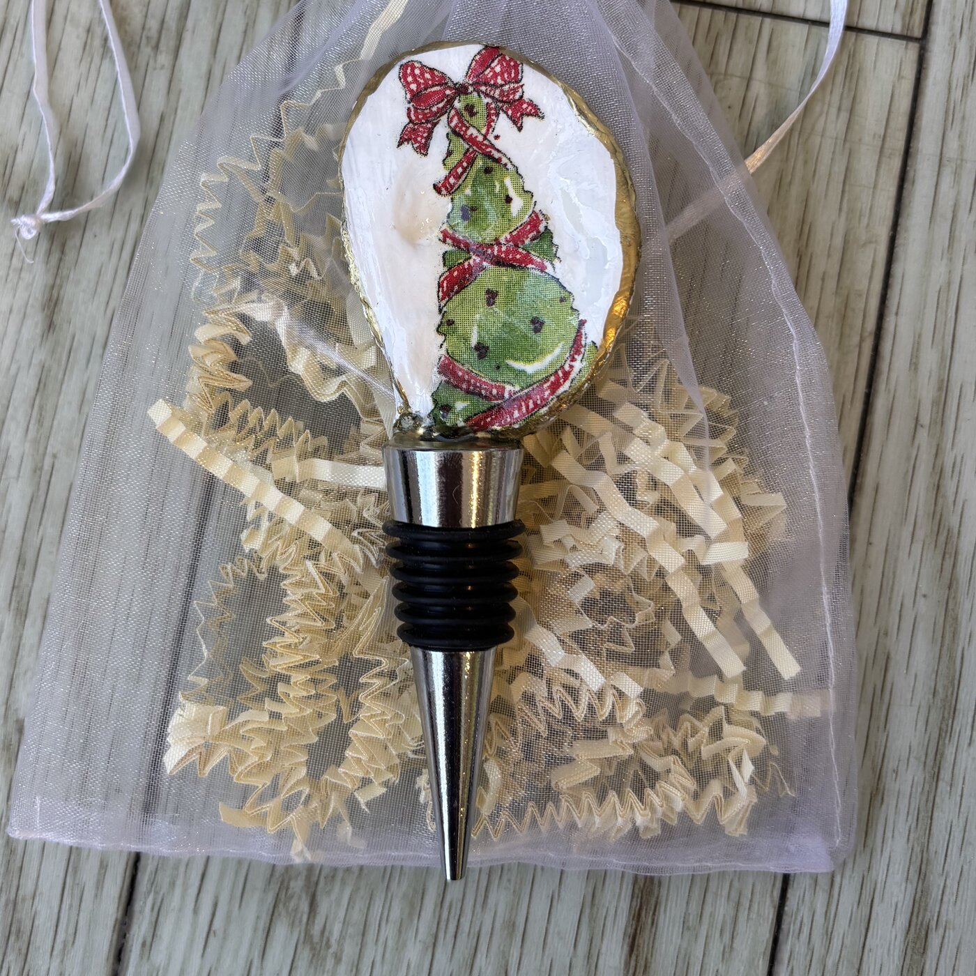 A wine bottle stopper topped with a gold-rimmed shell showing a ribbon-wrapped Christmas tree. Comes in a gift bag.