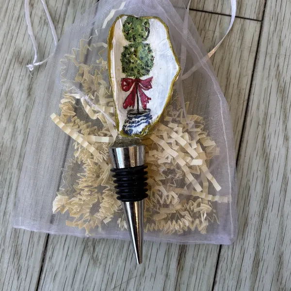 A wine bottle stopper topped with a gold-rimmed shell showing a topiary tree with a red bow. Comes in a gift bag.